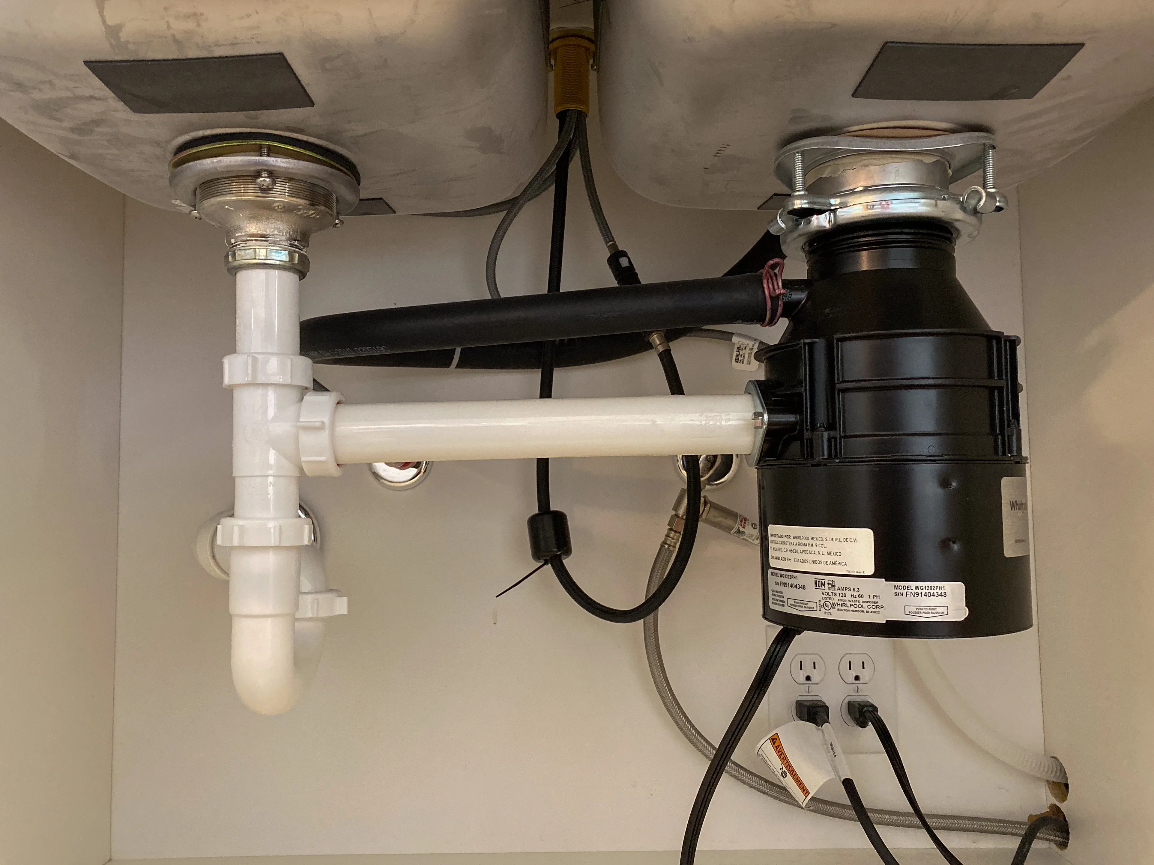 Under-sink view of a garbage disposal unit connected to white PVC drain pipes, showing the plumbing hookup for inspection and maintenance.